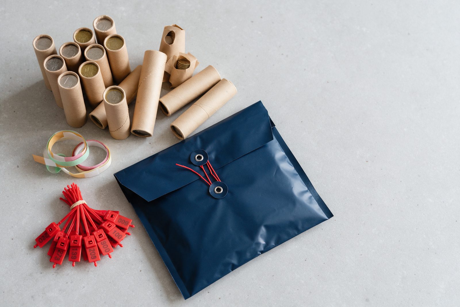 Tubes de pièces en papier kraft, bracelets de papier de couleur, scellés plastiques rouges numérotés et pochette fermée par un cordon, sur un plan de travail gris